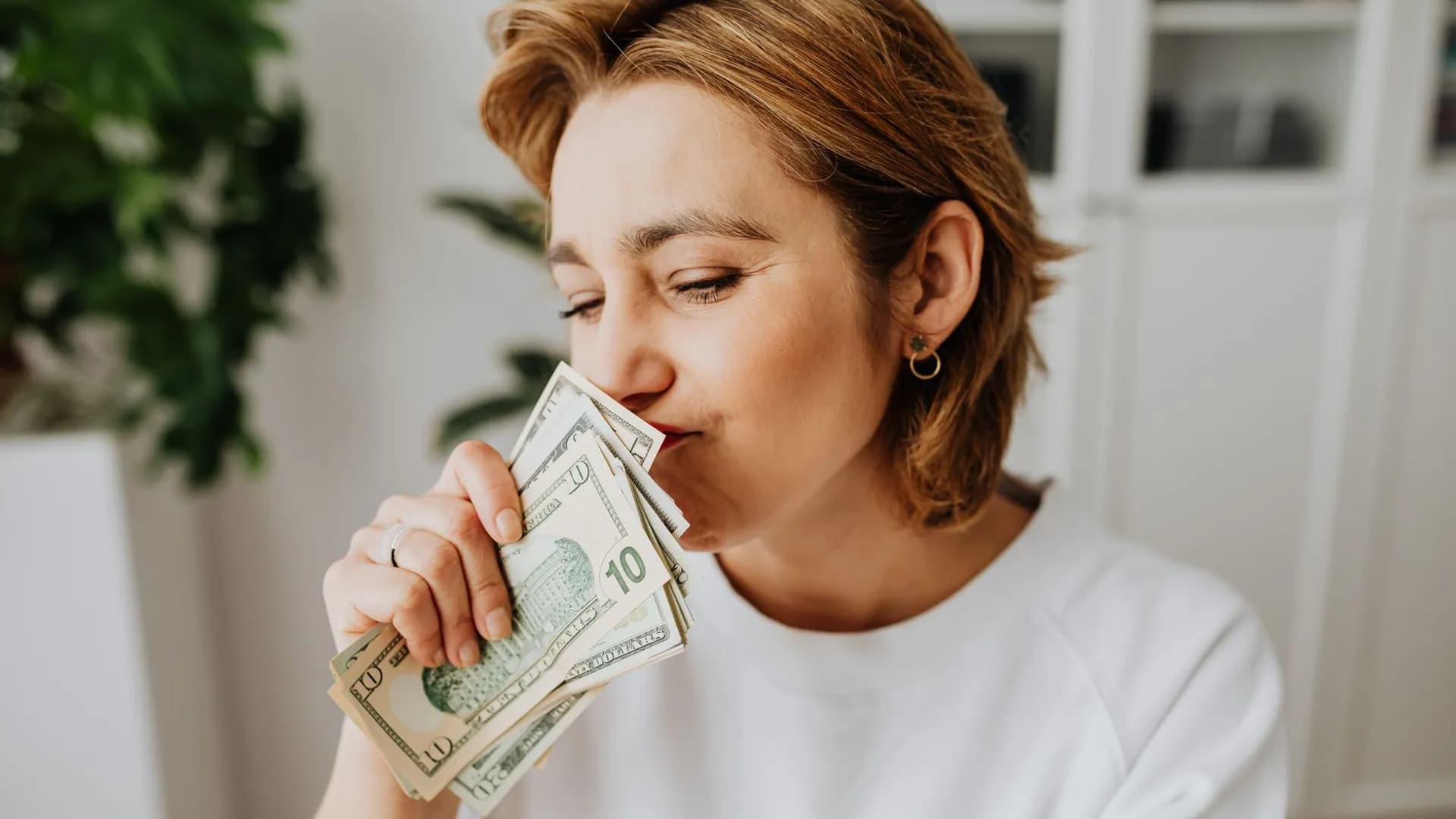 A woman joyfully smelling a handful of US dollar bills indoors.
