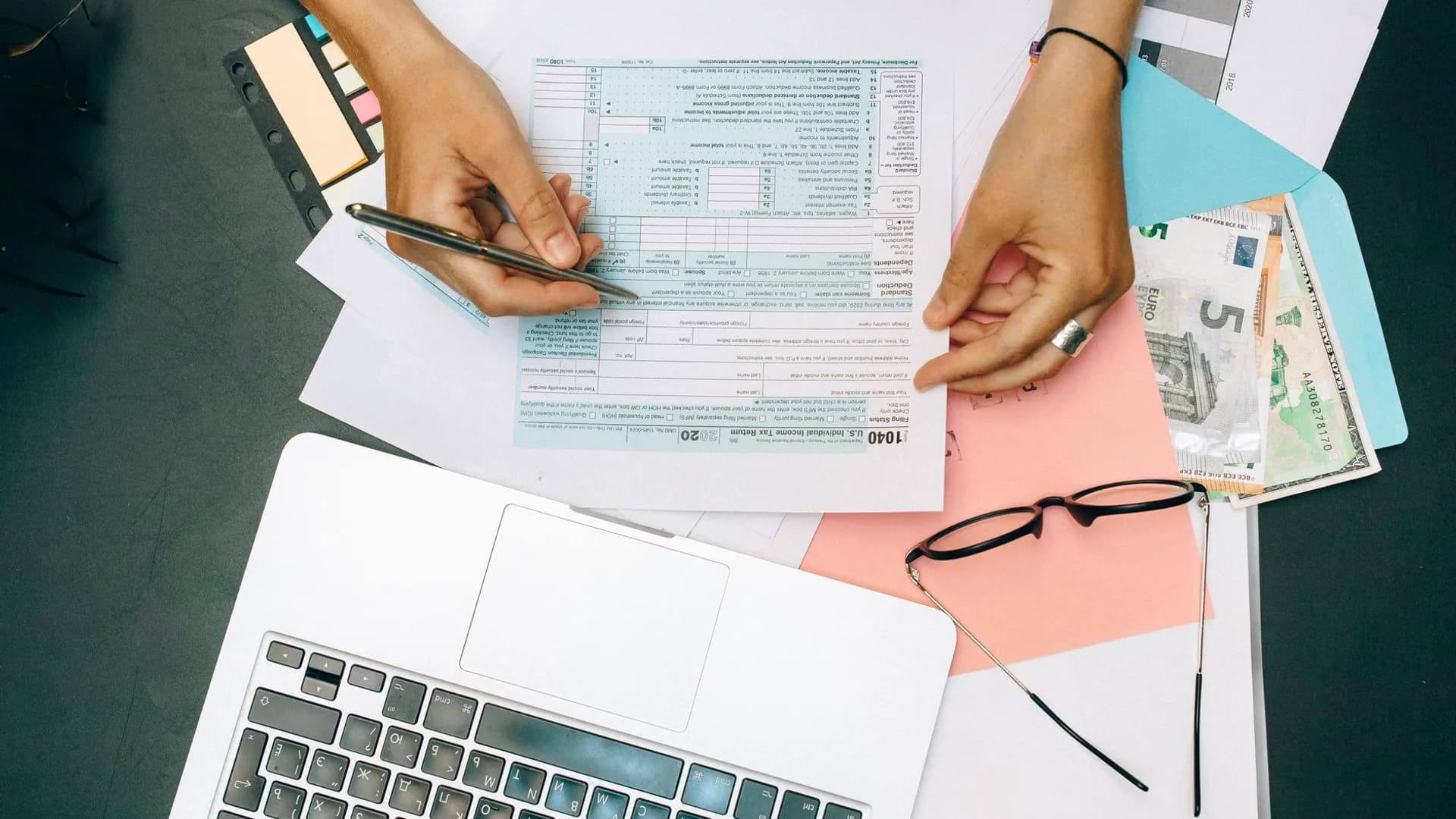 Hands writing on tax documents with laptop, glasses, and currency on desk.