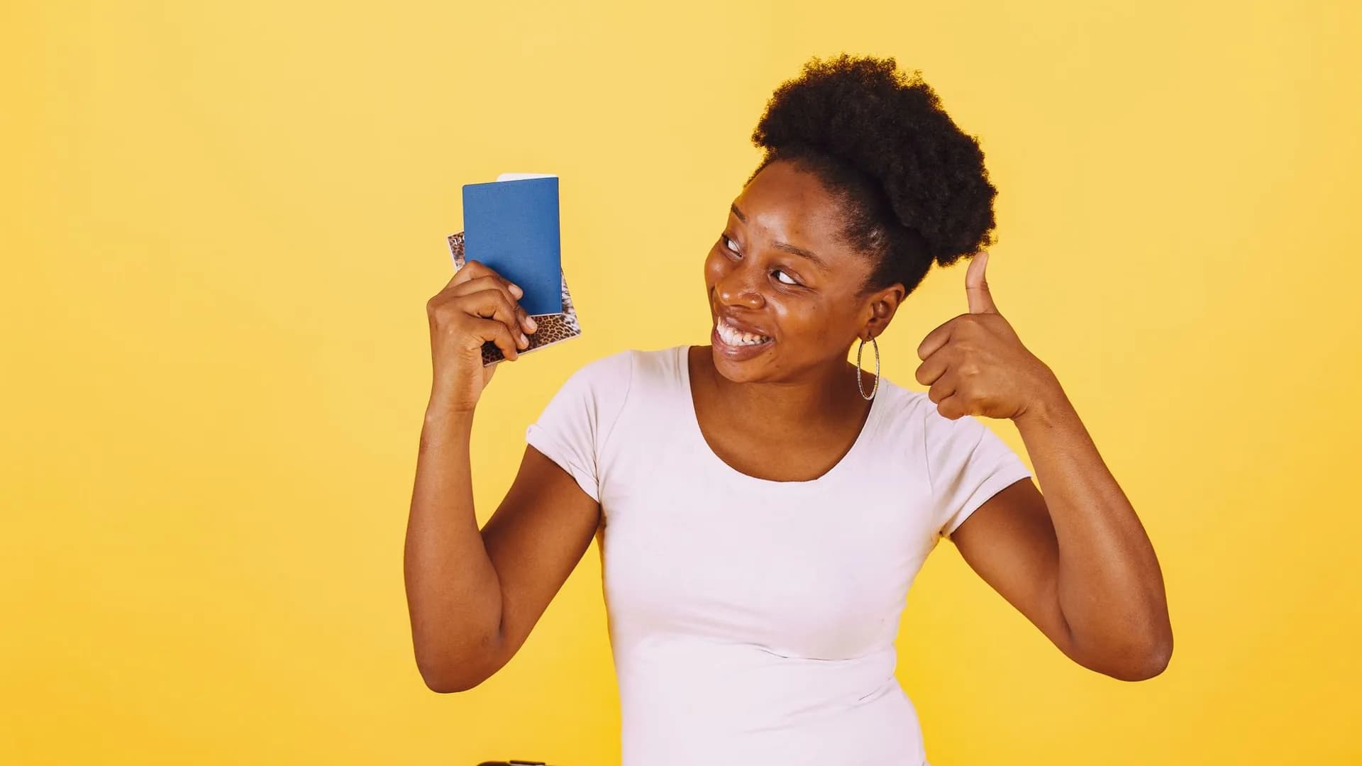 Happy woman holding passport with thumbs up, standing against yellow background, ready for travel.