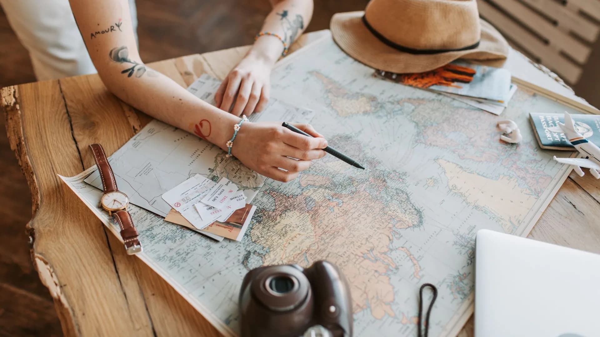 Tattooed woman planning a trip with a world map, passports, and travel essentials on a wooden table.