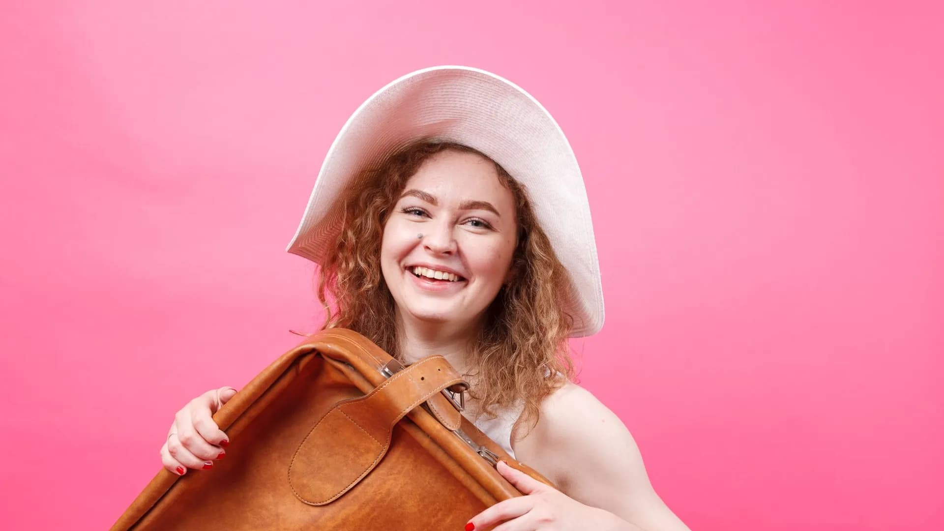 Happy young woman holding a suitcase, wearing a hat, smiling in a pink studio setting.