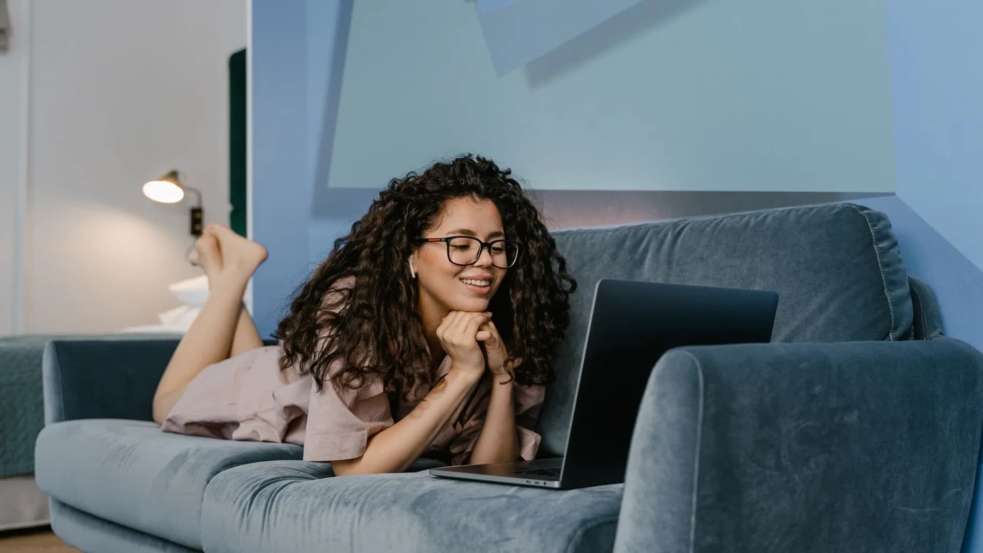 Happy woman with curly hair video calling on laptop, lying on sofa in cozy living room.