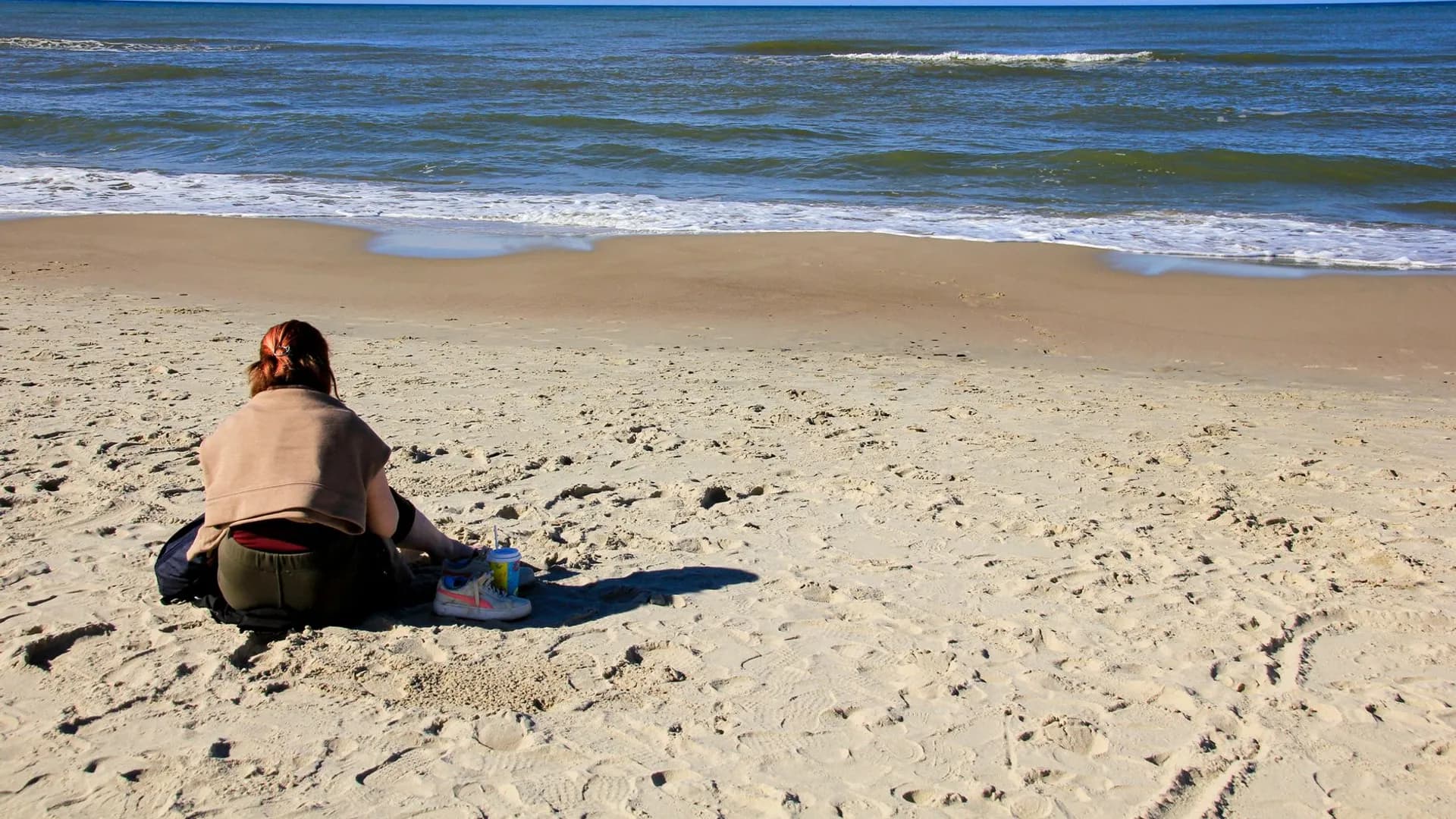A woman sits peacefully on a sandy beach gazing at the ocean waves, capturing a serene moment of solitude.