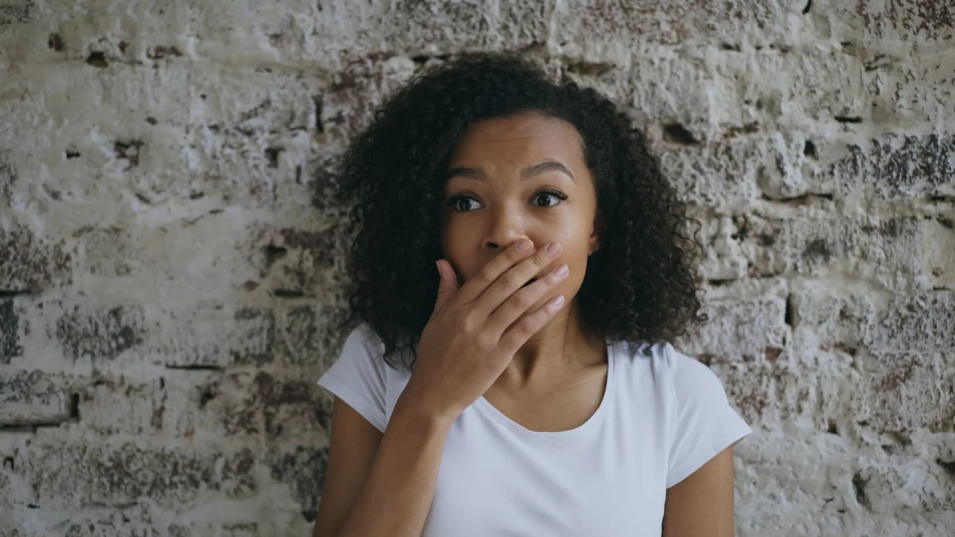 Young woman covering her mouth in shock against a rustic brick wall background.
