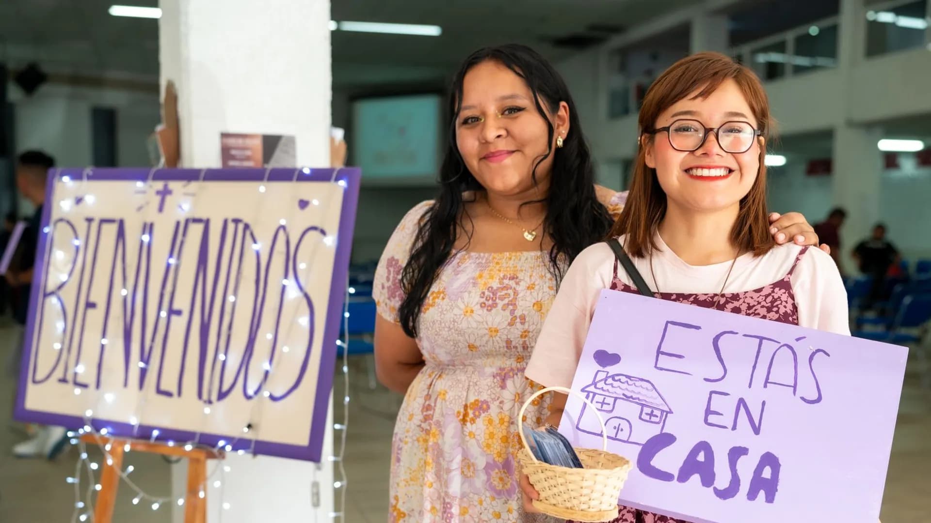 Two women warmly greet visitors at a community event in Ciudad de México with signs saying 'Bienvenidos' and 'Estás en Casa'.