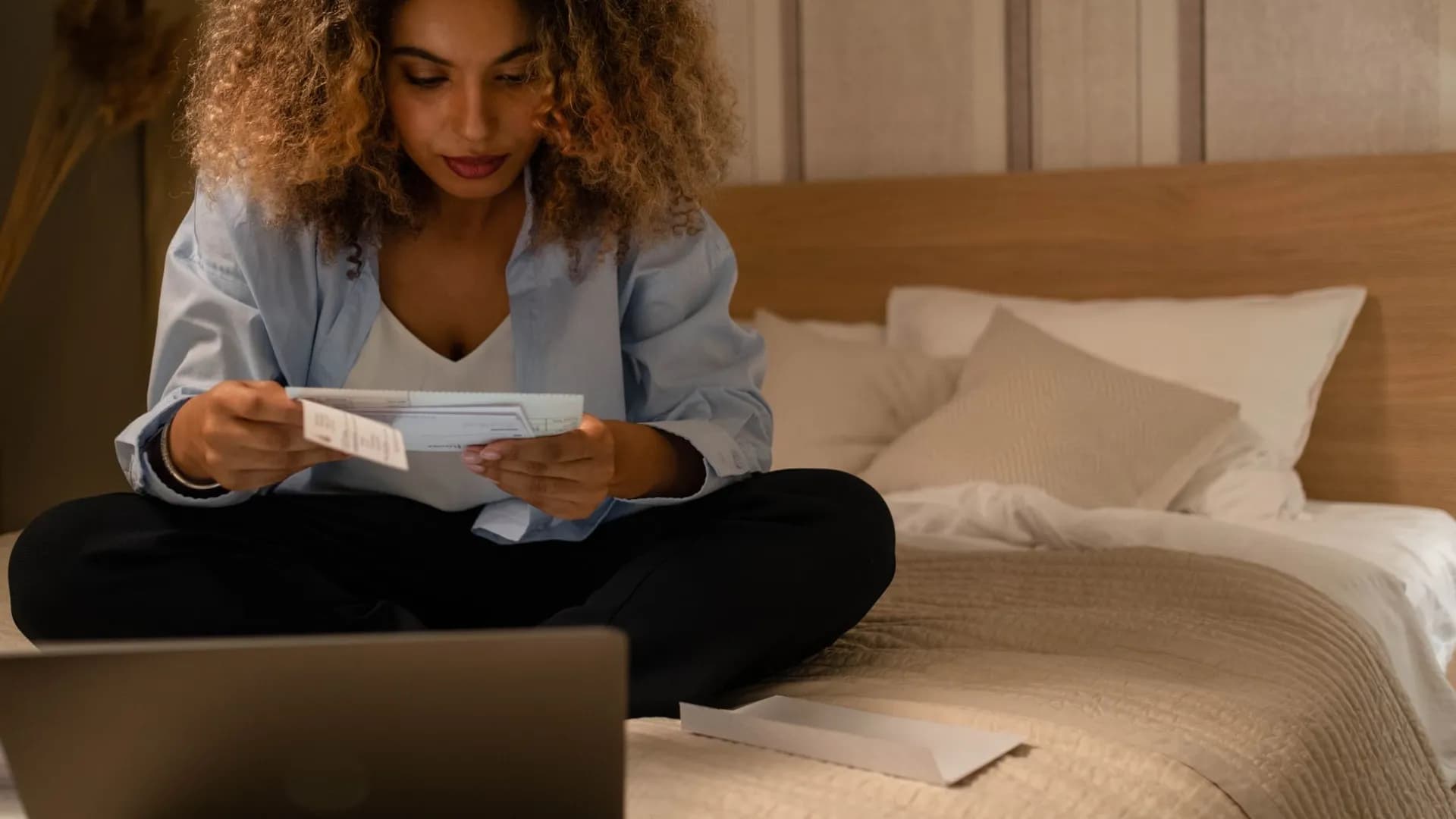 Woman sitting on bed reading documents with laptop, highlighting home office setting.