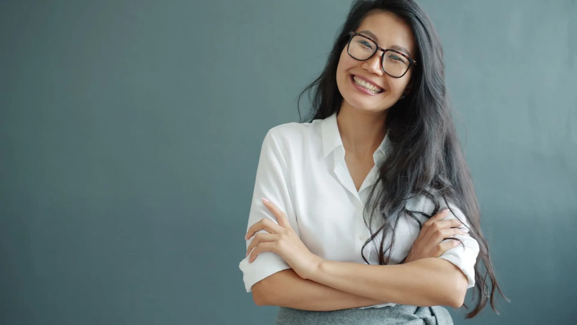 Portrait of a confident young woman smiling with folded arms, wearing glasses and a white shirt.