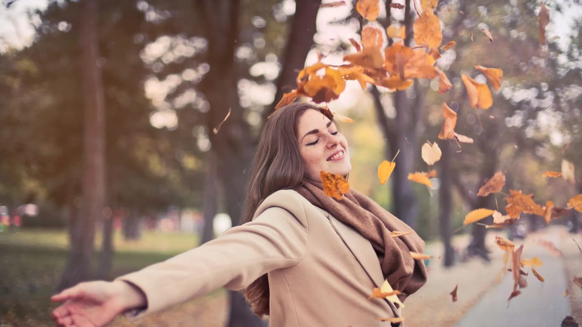 Young woman in a beige coat joyfully throwing autumn leaves in a park.
