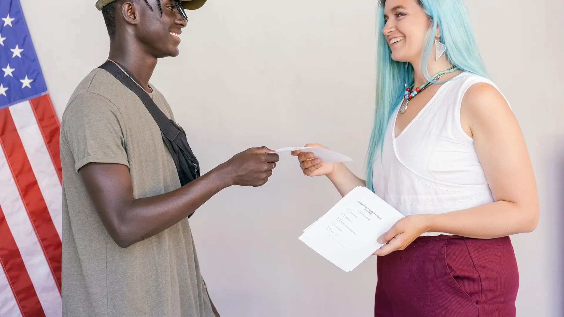 A diverse group of young adults exchanging papers indoors with an American flag in the background.