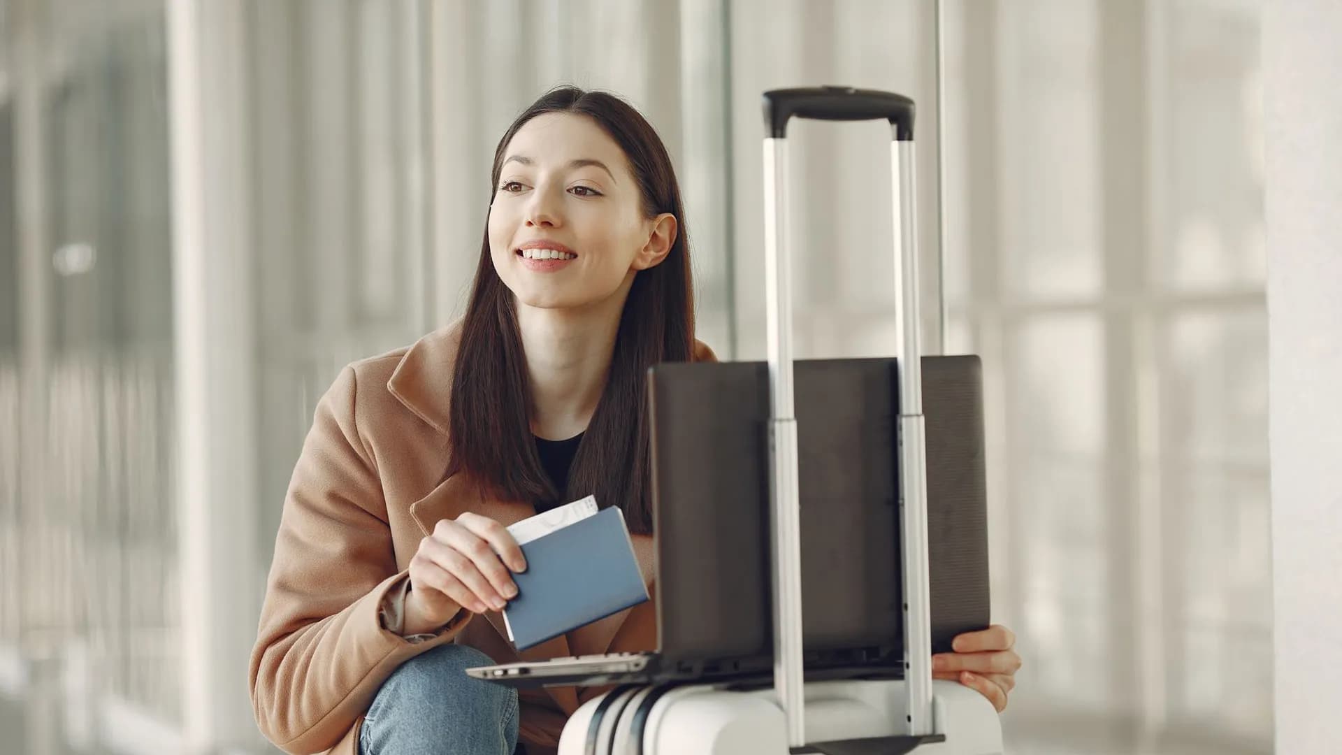 Smiling adult ethnic female traveler in trendy coat holding passport and using laptop on luggage in airport corridor for checking ticket for correctness