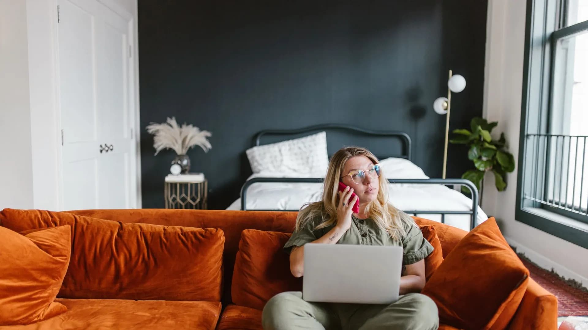 Woman sits on a vibrant sofa working on her laptop, talking on the phone in a cozy home setting.