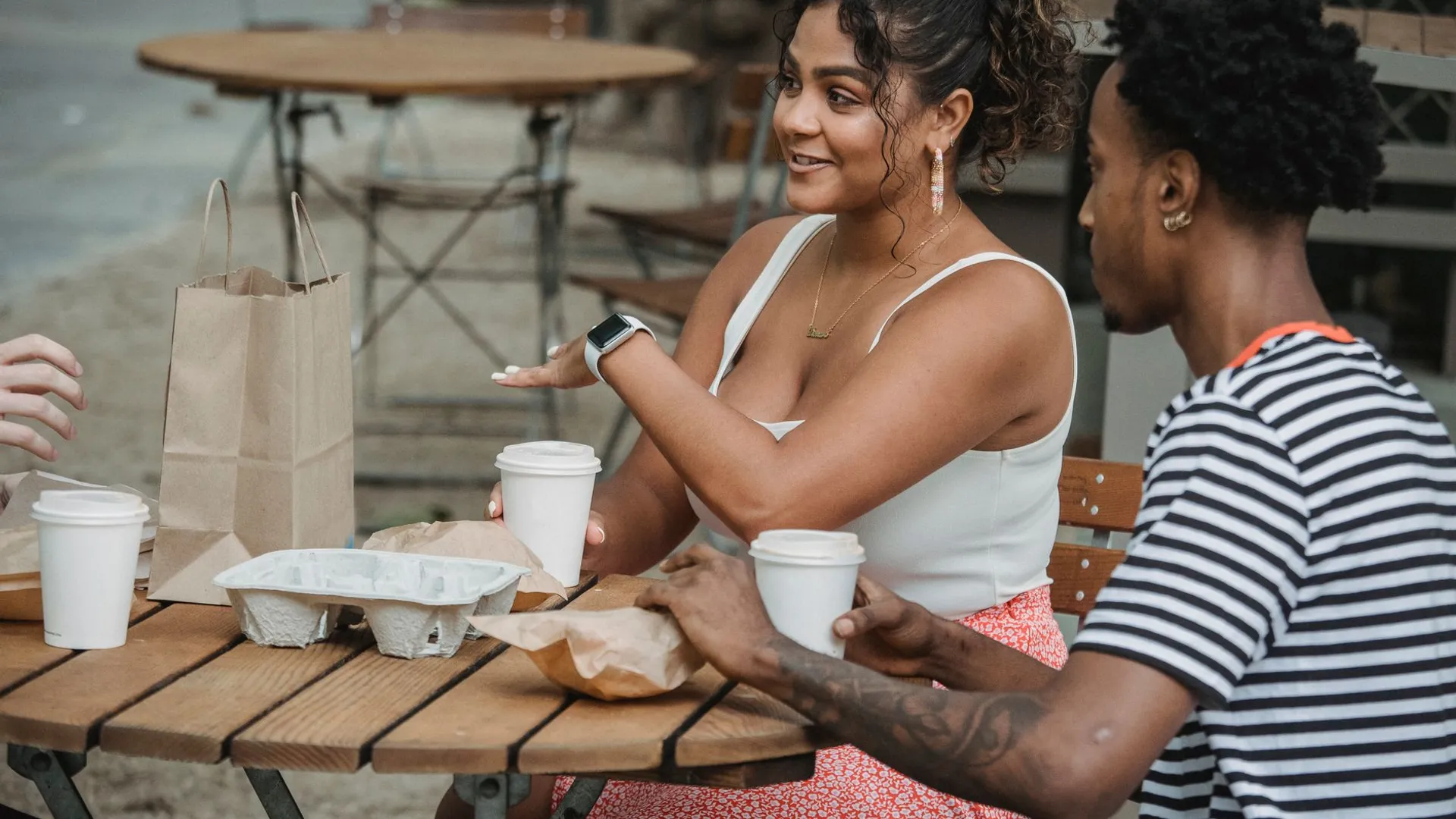 Side view of young happy Hispanic woman with multiracial friends chatting while resting in outdoor cafe with takeaway coffee and pastries