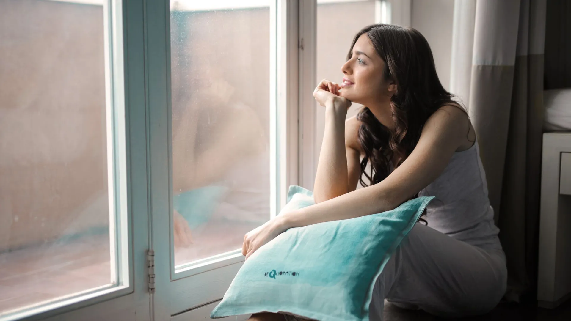 A thoughtful woman gazing outside while sitting by a window with a pillow.