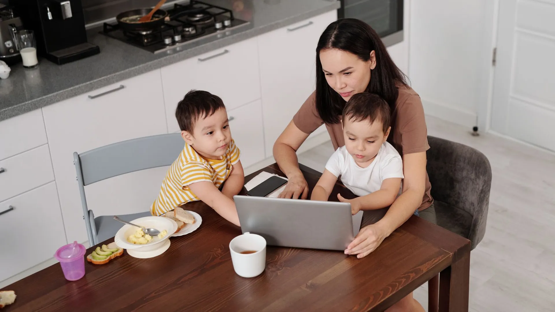 A mother multitasks on her laptop with her two young sons in a modern kitchen setting.