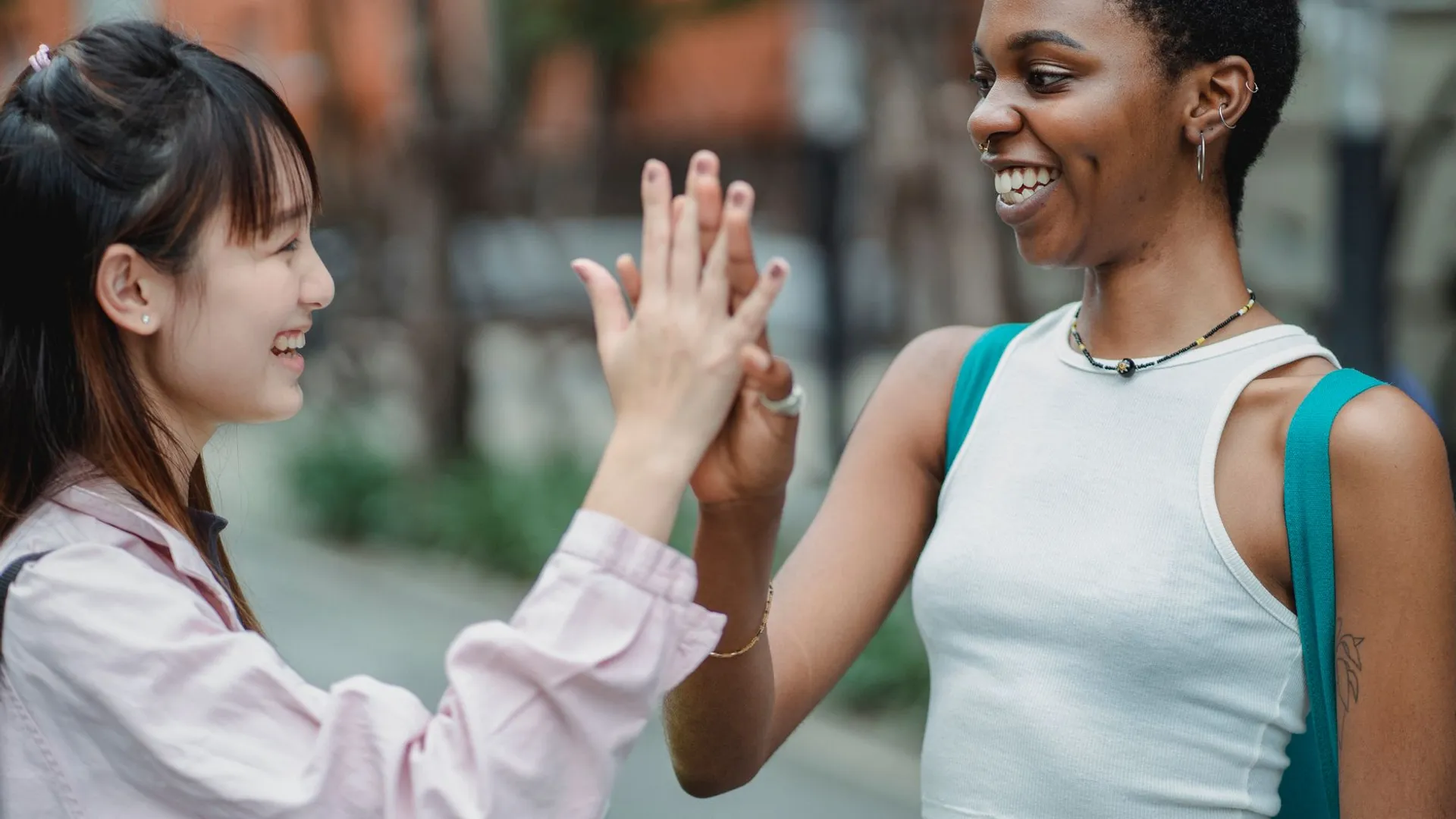 Smiling young multiethnic girlfriends wearing casual clothes while standing in middle of road and giving high five in summer day while looking at each other