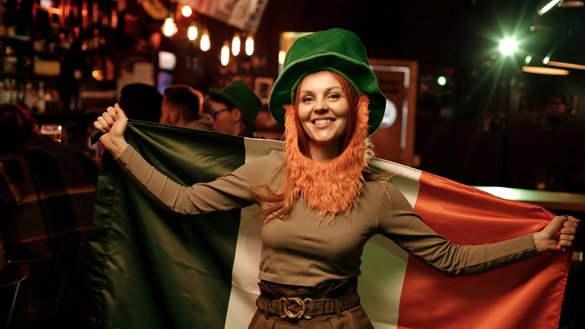 Smiling woman in green hat holding Irish flag at St. Patrick's Day celebration in a lively pub.