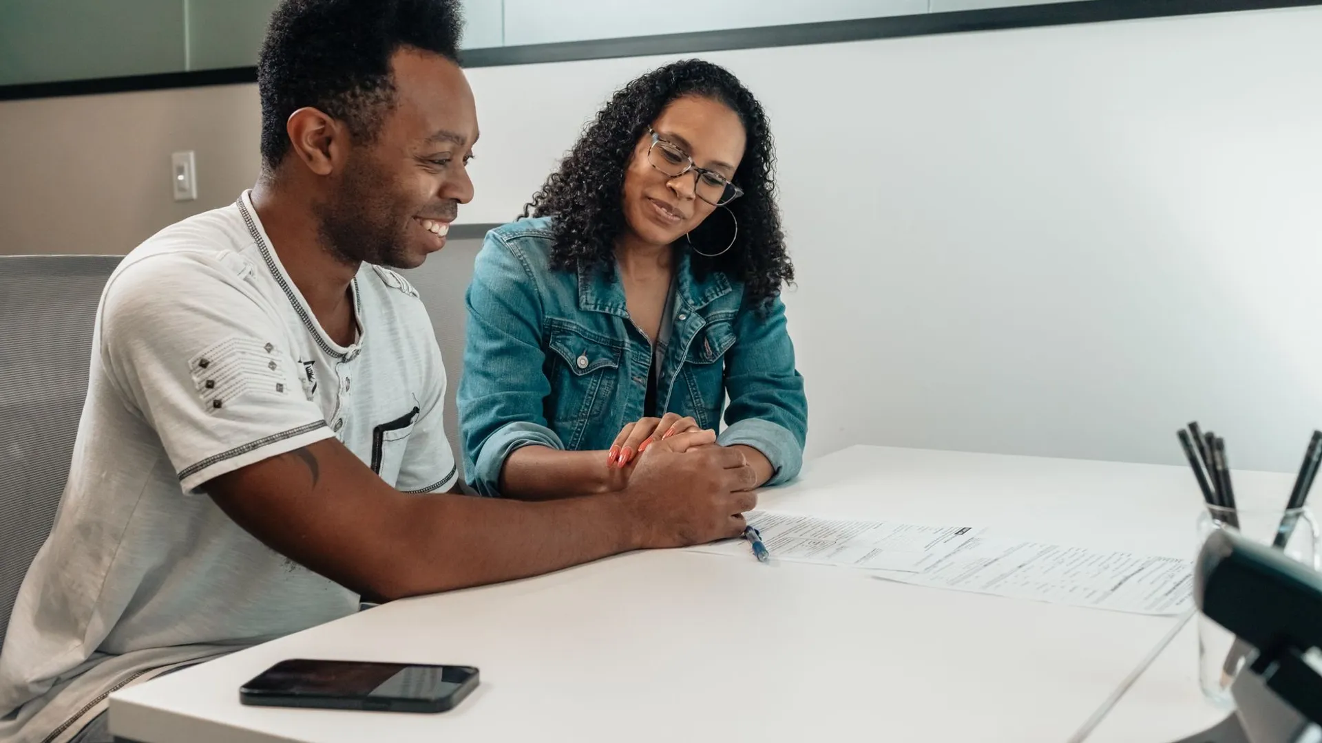 A couple sits at a white table, happily reviewing documents and holding hands in an office setting.