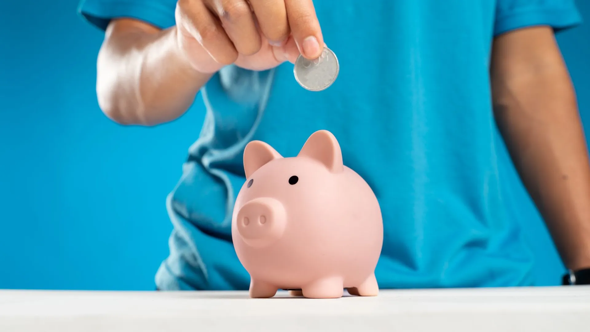 A close-up of an adult's hand dropping a coin into a piggy bank, symbolizing savings and investment.