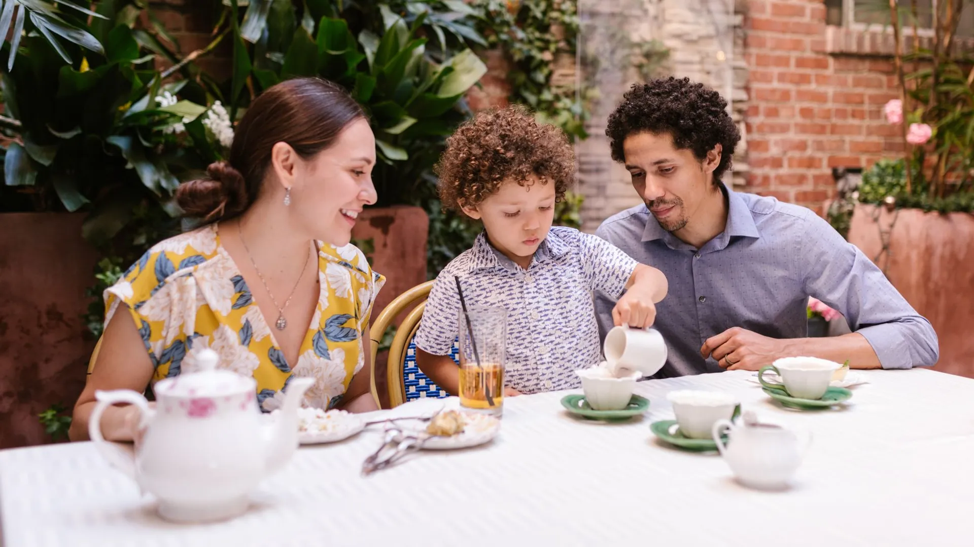Family having a joyful tea time at an outdoor café. A young boy pours tea with his parents nearby, surrounded by lush plants.