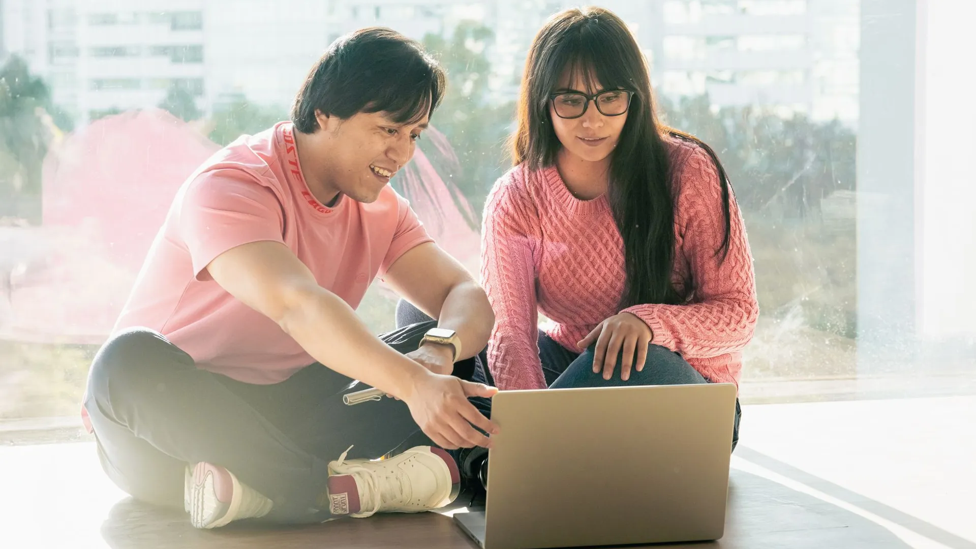 Two young adults in casual attire working together on a laptop by a window.